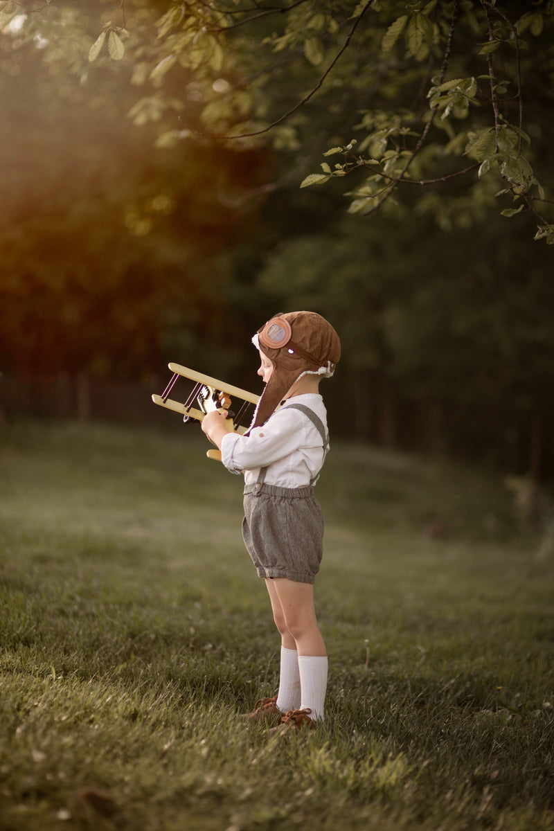 Child in green suspender shorts holding a toy airplane in a natural setting with trees and grass.
