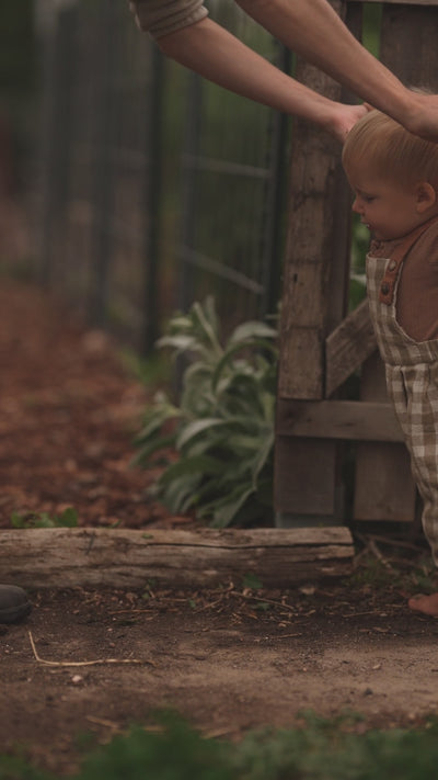 Baby learning to walk in beige gingham linen overalls, showcasing the comfortable and breathable fabric, adjustable straps for a perfect fit, and unfoldable leg cuffs designed to accommodate growth.