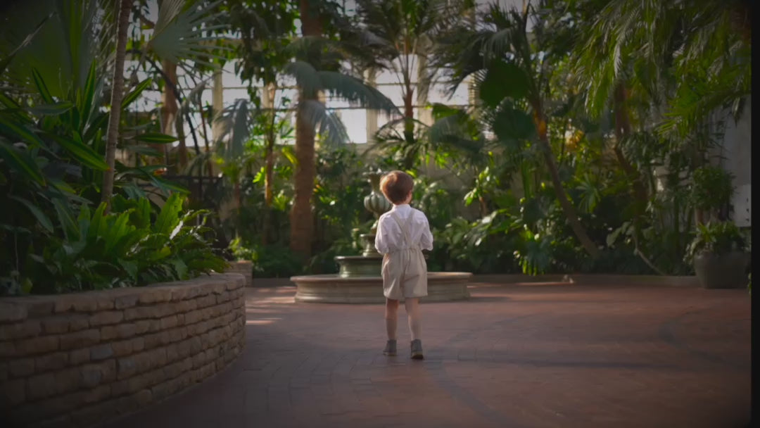 A little boy in a beige stripes Grow-With-Me Romper walking through a greenhouse surrounded by various plants and flowers, showcasing adjustable straps, comfortable fabric, and timeless design.