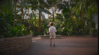 A little boy in a beige stripes Grow-With-Me Romper walking through a greenhouse surrounded by various plants and flowers, showcasing adjustable straps, comfortable fabric, and timeless design.