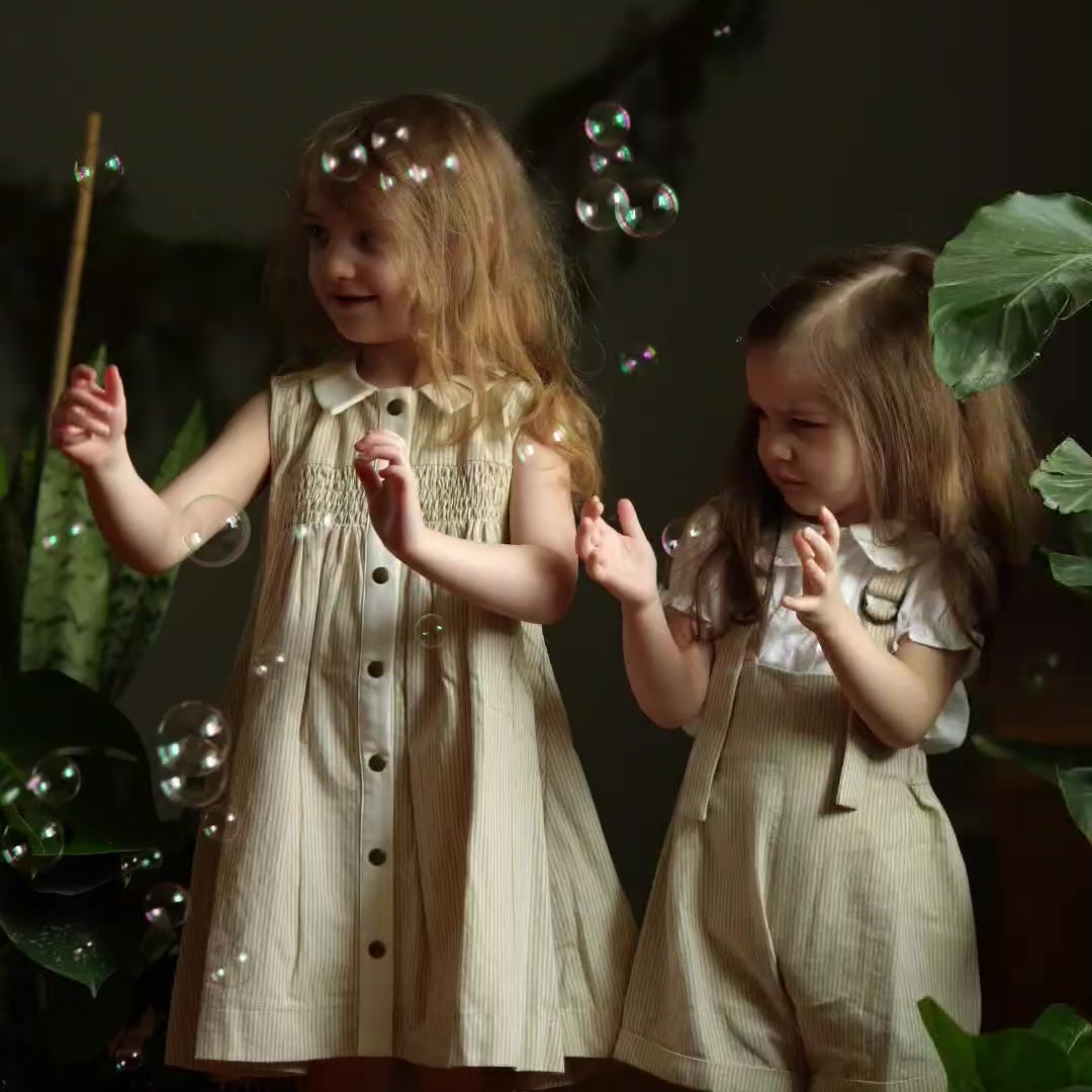 Video of a girl and her little sister both wearing Beige Stripes Dress with white contrasting collar and button placket, joyfully playing with soap bubbles, capturing moments of fun and elegance in sustainable children's wear.