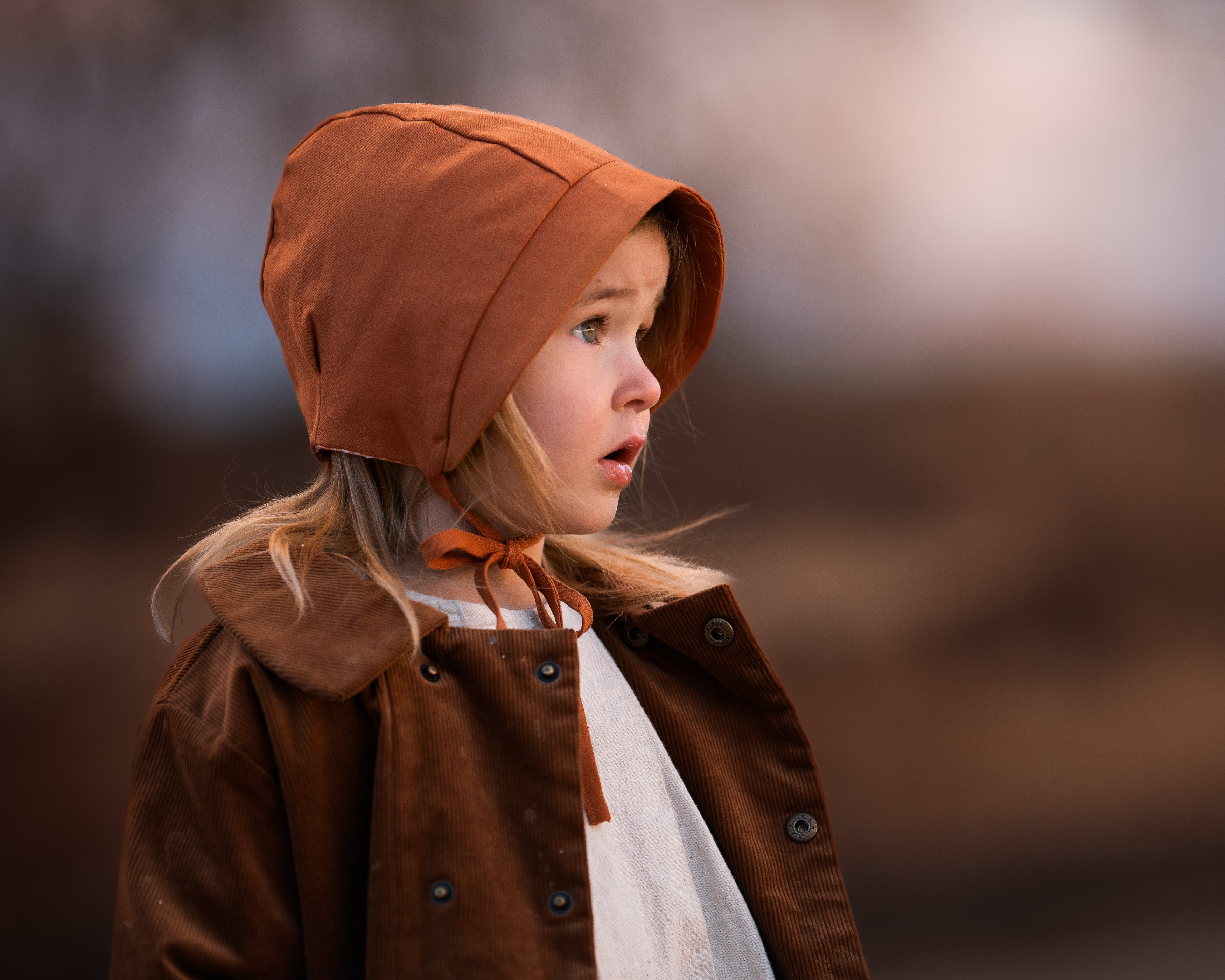 Side view of the Burnt Orange Bonnet on a girl model - Showcasing the pleats on each side for an adjustable fit, made from soft linen fabric that keeps little heads cool and protected during summer.