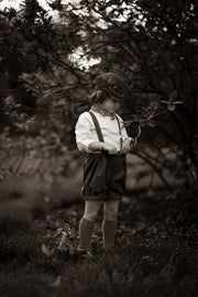 Black and white photo of a child in suspenders and shorts standing in a natural setting.