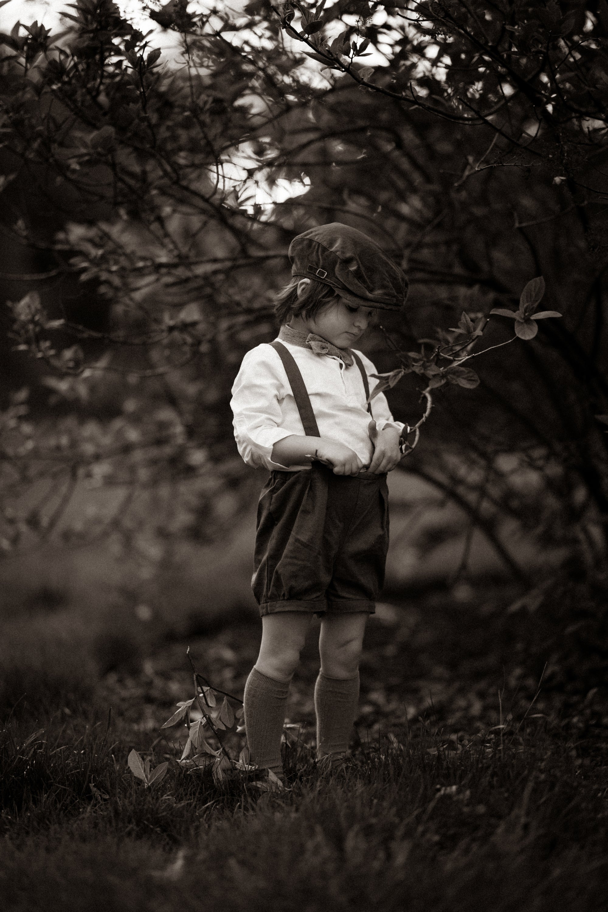 Black and white photo of a child in suspenders and shorts standing in a natural setting.