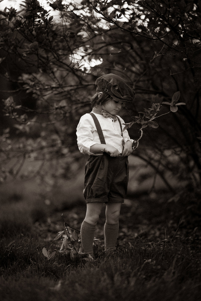 Black and white photo of a child in suspenders and shorts standing in a natural setting.
