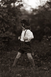 A model boy holding the adjustable straps of his black linen suspender shorts, emphasizing the customized fit and style, with a focus on his comfort and the details of his outfit in a natural setting.