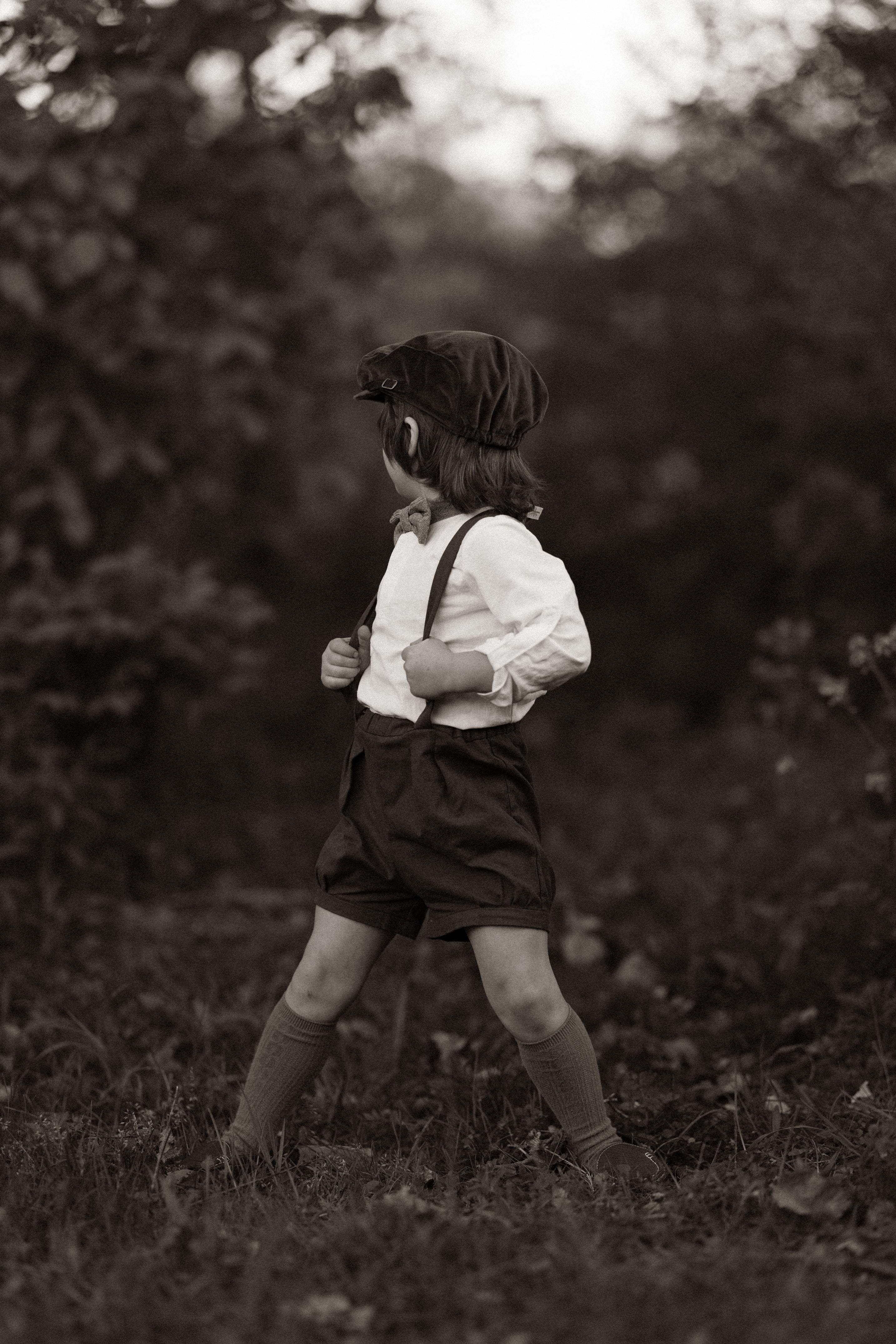 A model boy holding the adjustable straps of his black linen suspender shorts, emphasizing the customized fit and style, with a focus on his comfort and the details of his outfit in a natural setting.