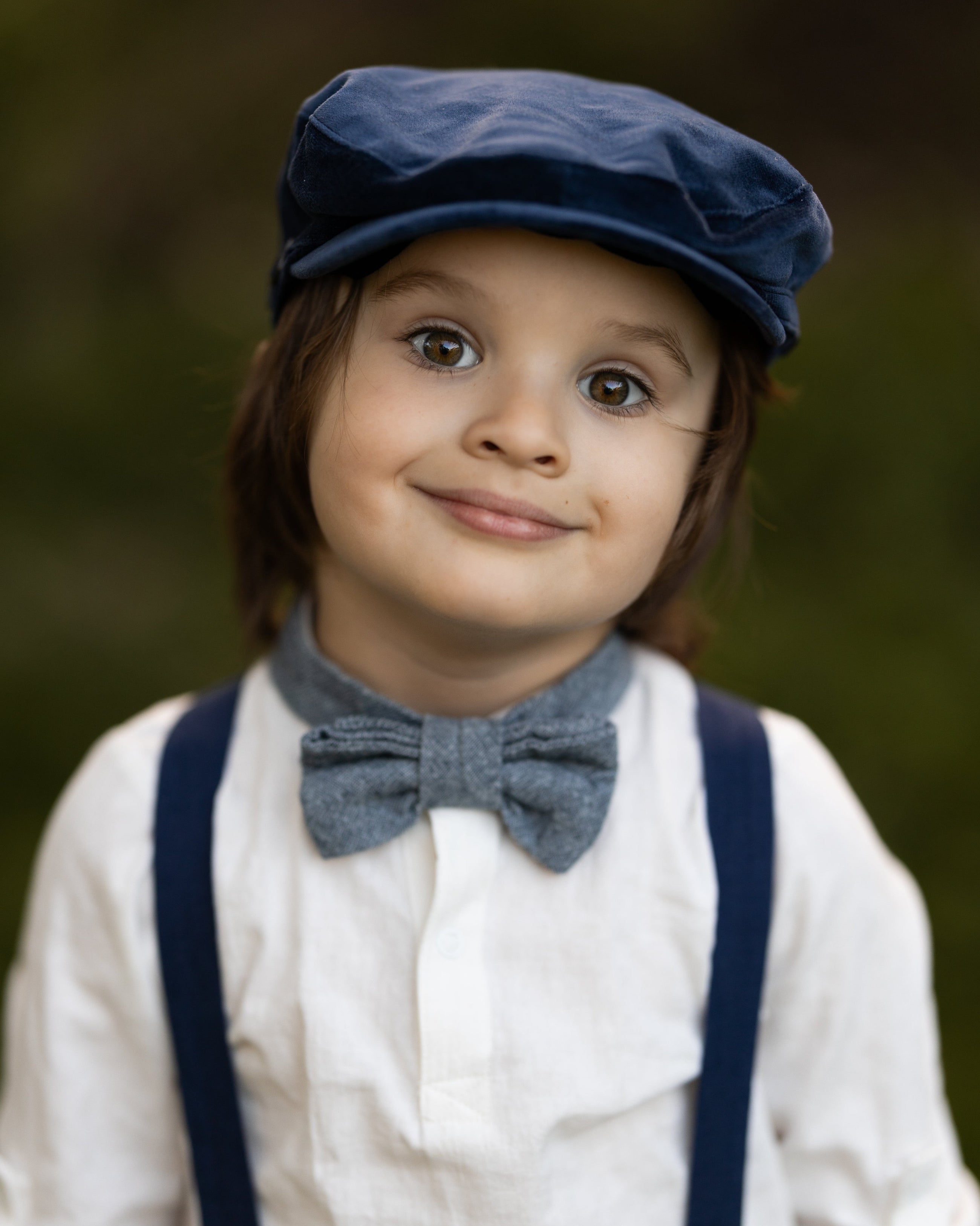 Child wearing a blue cap, suspenders, and bow tie against a blurred background