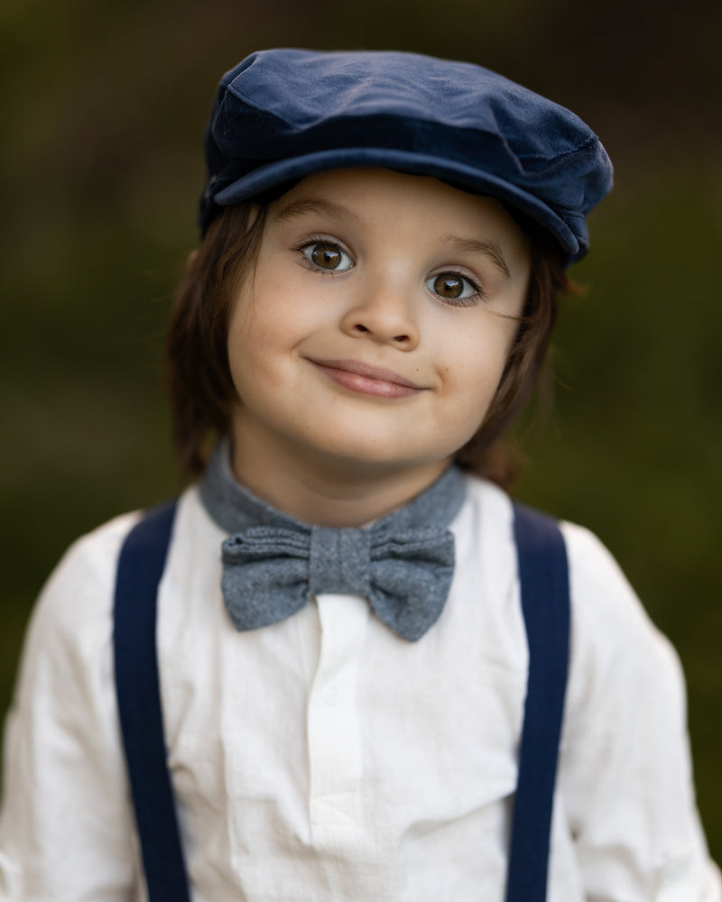 Child wearing a blue cap, suspenders, and bow tie against a blurred background