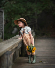 Girl in yellow gingham overalls and hat holding sunflowers on a wooden path with greenery in the background