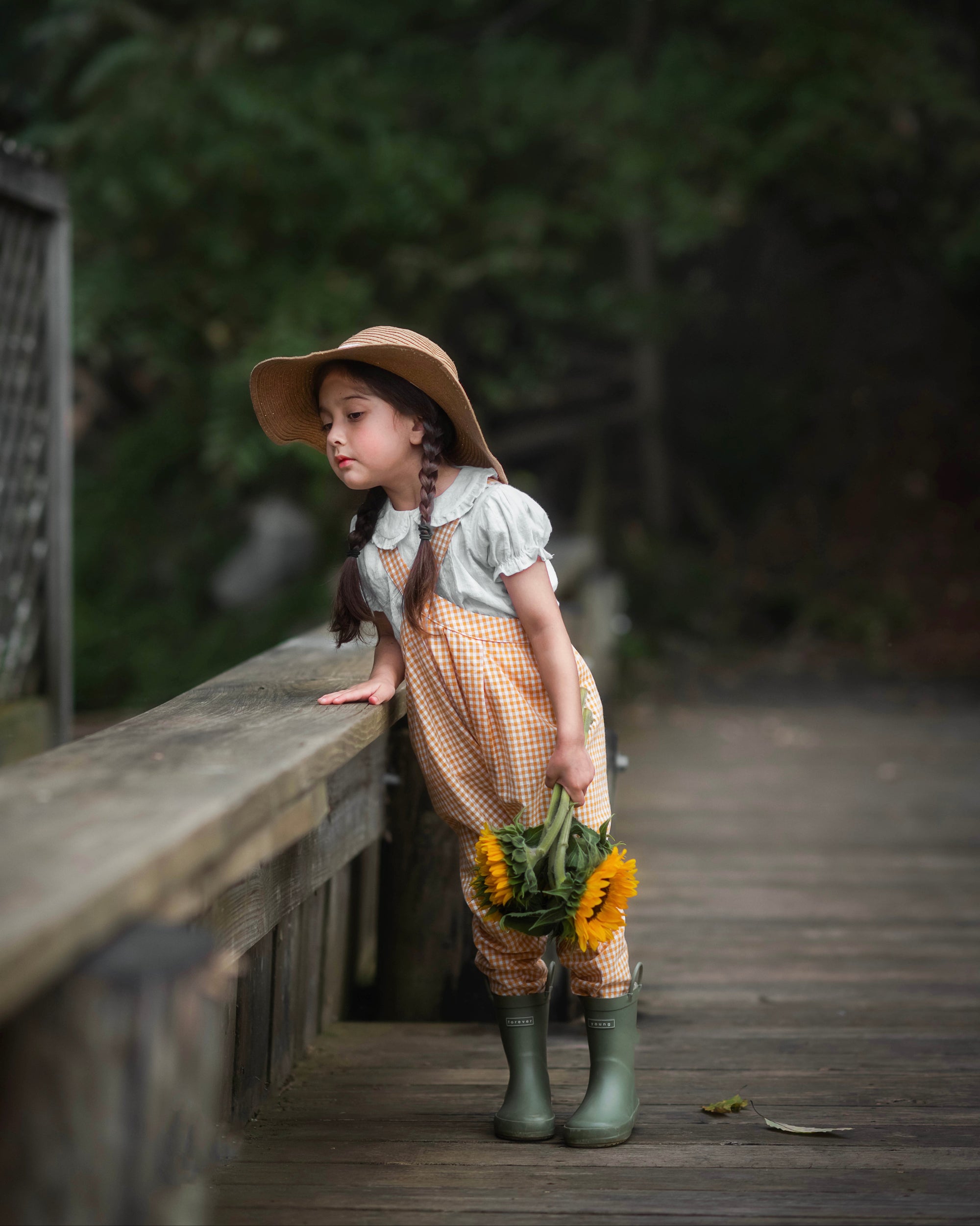 Girl in yellow gingham overalls and hat holding sunflowers on a wooden path with greenery in the background