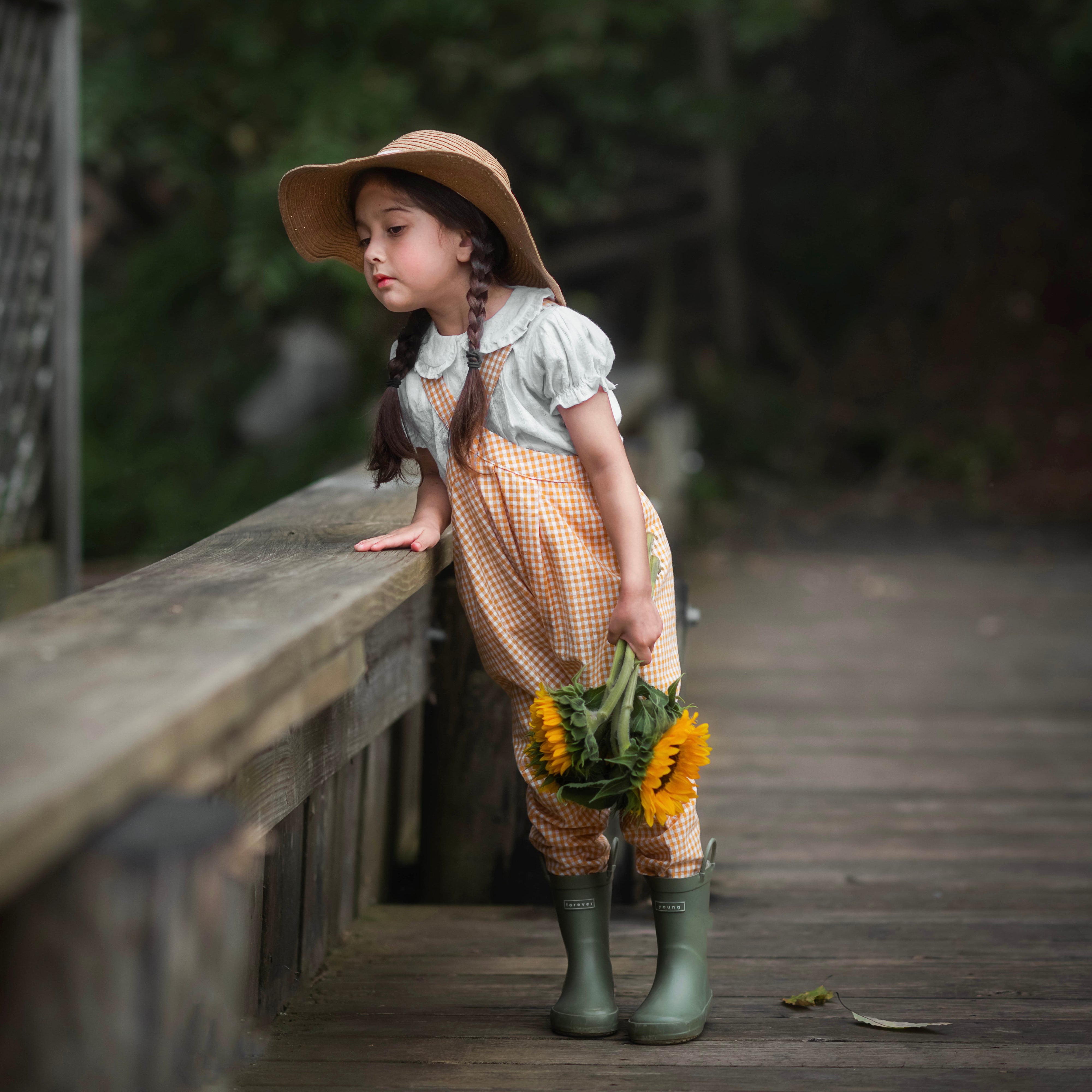 Girl in yellow gingham overalls and hat holding sunflowers on a wooden path with greenery in the background