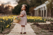 Young girl in a pink dress holding a basket of flowers in a garden with yellow flowers.
