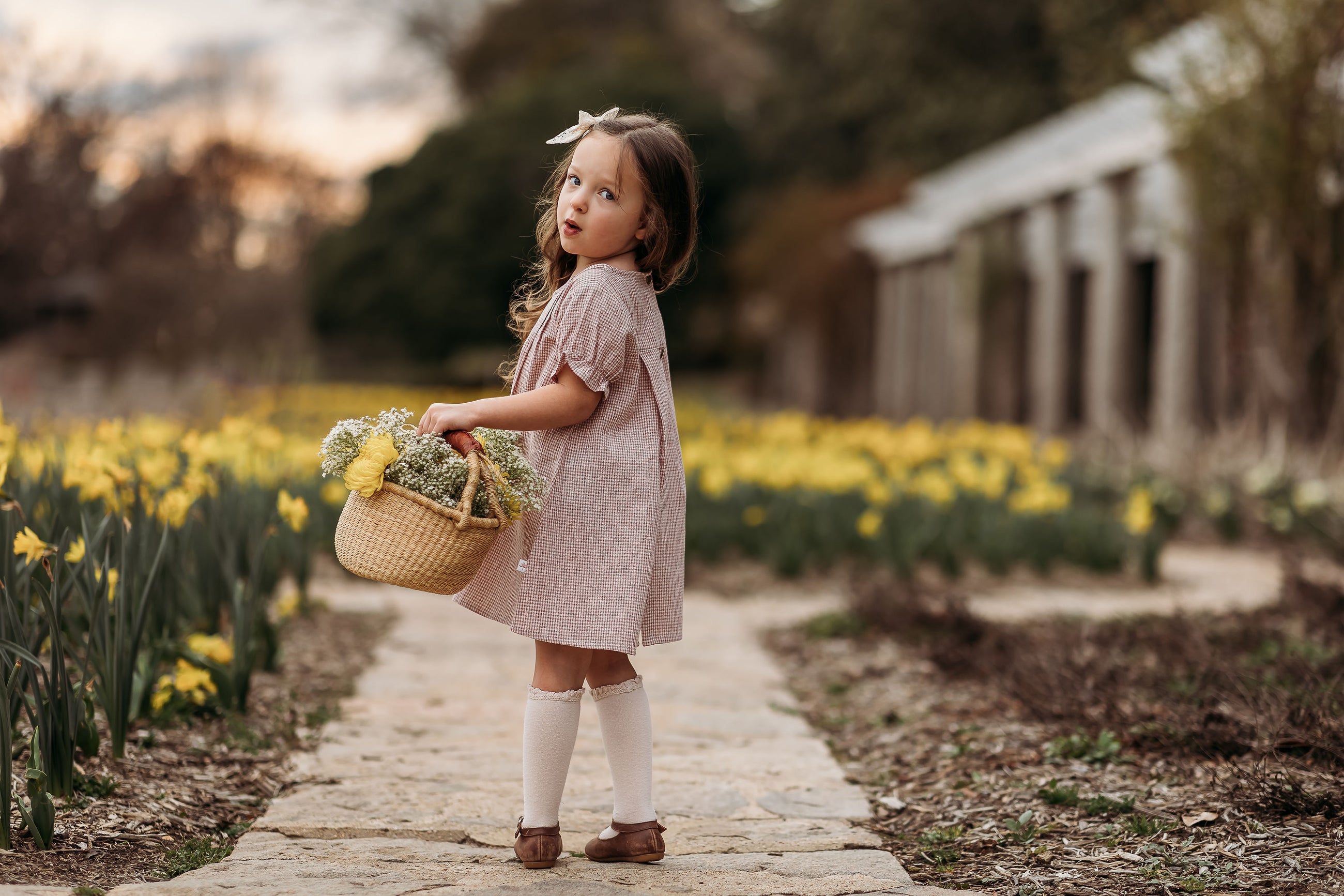 Young girl in a pink dress holding a basket of flowers in a garden with yellow flowers.