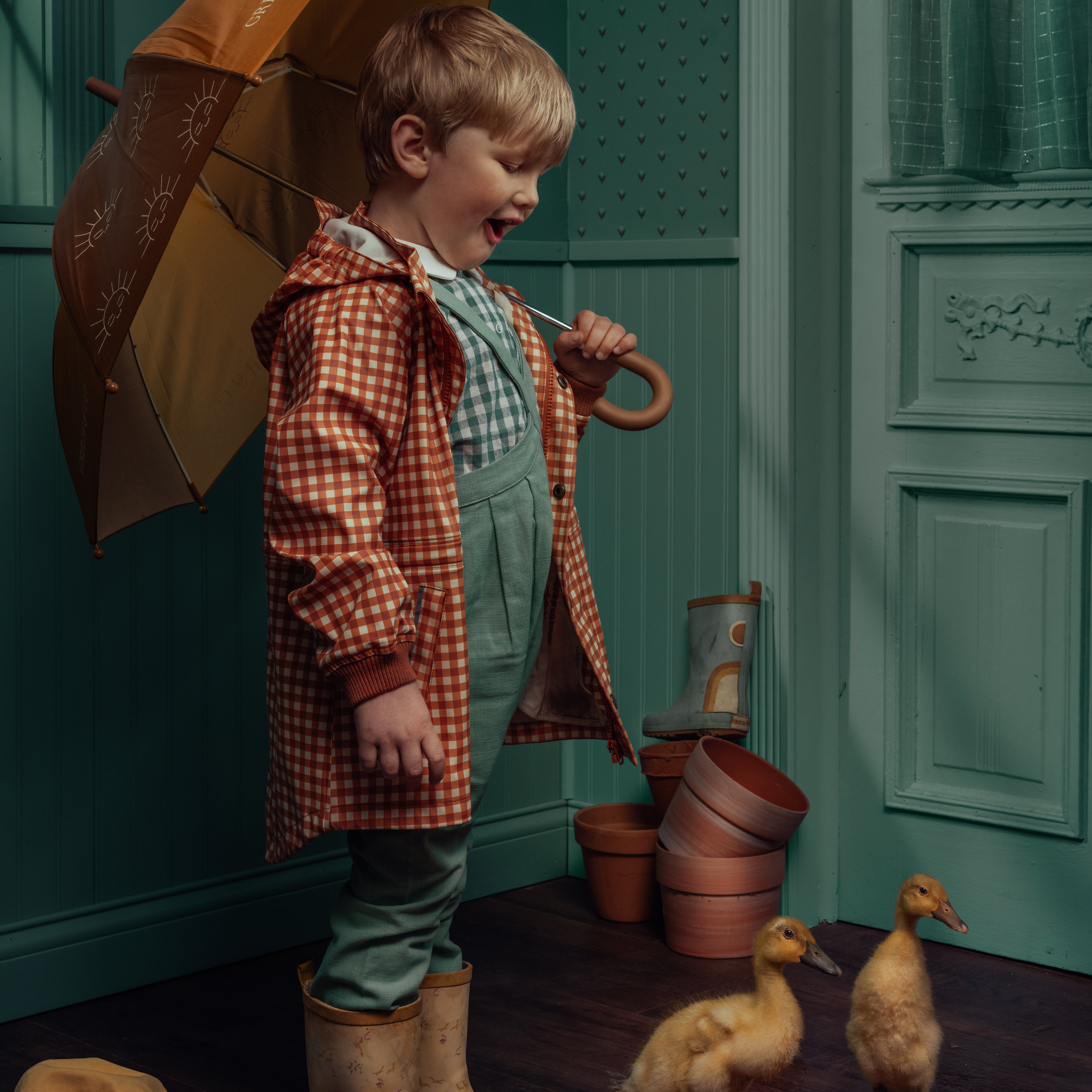 Child in a checkered raincoat and boots holding an umbrella, standing next to toy ducks and pots in a room.