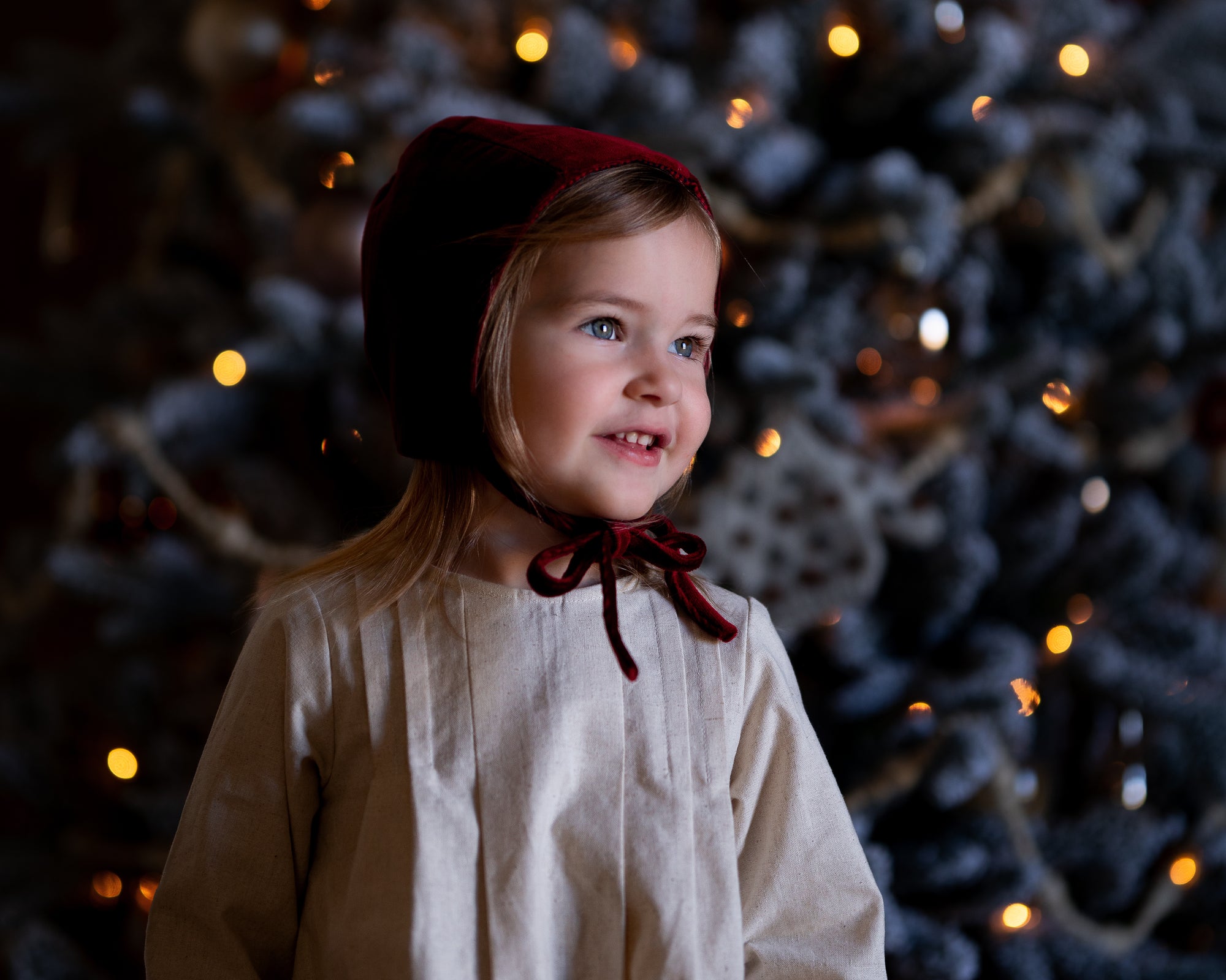 Child wearing a red bonnet in front of a decorated Christmas tree with lights.